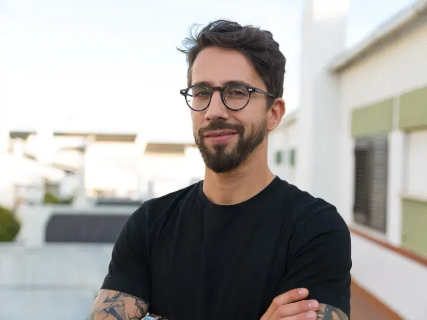 Lächelnder Mann mit Brille, der mit verschränkten Armen auf einer Dachterrasse steht.
